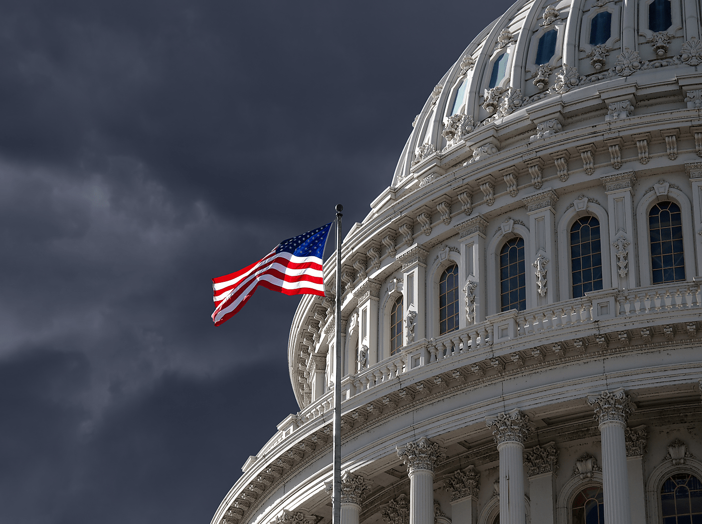 An American flag waves in front of the Capitol with a stormy sky